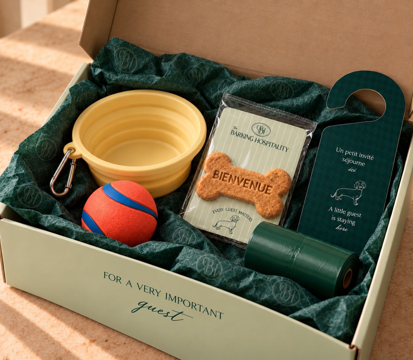 Coffret ouvert sur un papier de soie vert sarcelle monogrammé : accroche-porte, biscuit «&nbsp;Bienvenue&nbsp;», gamelle pliable, sacs de propreté et balle de jeu